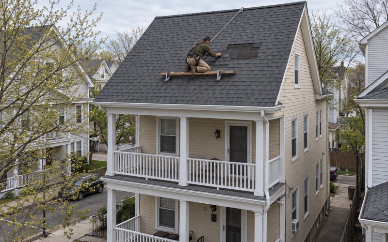 A roofer sliding replacement shingles into a gap where wind took several off a Perth Amboy two-family roof.